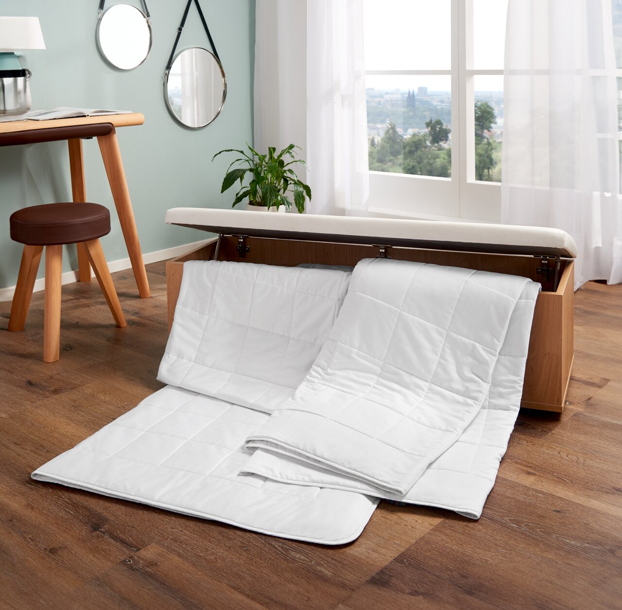 Wildseidendecke Superlight in einem modernen Raum, teilweise aus einer Holztruhe herausgezogen, mit Blick auf ein Fenster mit Vorhängen.