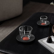 Kaffeeuntertasse Manufacture mit transparentem Glas, seitliche Perspektive auf einem dunklen Tisch.
