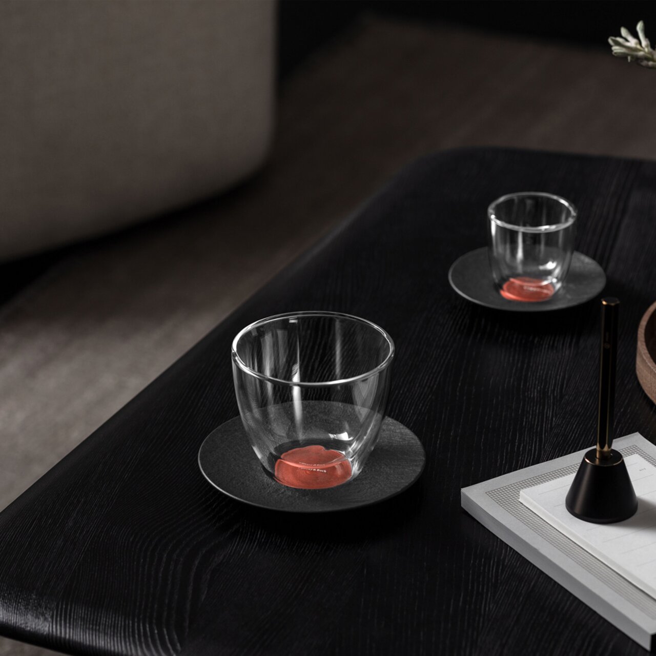 Kaffeeuntertasse Manufacture mit transparentem Glas, seitliche Perspektive auf einem dunklen Tisch.