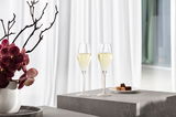 Zwei Champagnergläser mit Sekt gefüllt, auf einem grauen Podest stehend, mit einem Teller Pralinen daneben und einer Vase mit Orchideen im Hintergrund, seitliche Perspektive.