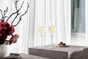Twee met mousserende wijn gevulde champagneglazen, staande op een grijs voetstuk, met een schaal pralines ernaast en een vaas met orchideeën op de achtergrond, zijaanzicht.