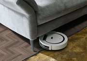 Ein graues Ecksofa mit einem Saugroboter, der darunter hervorkommt. Perspektive von oben auf das Sofa und den Boden.