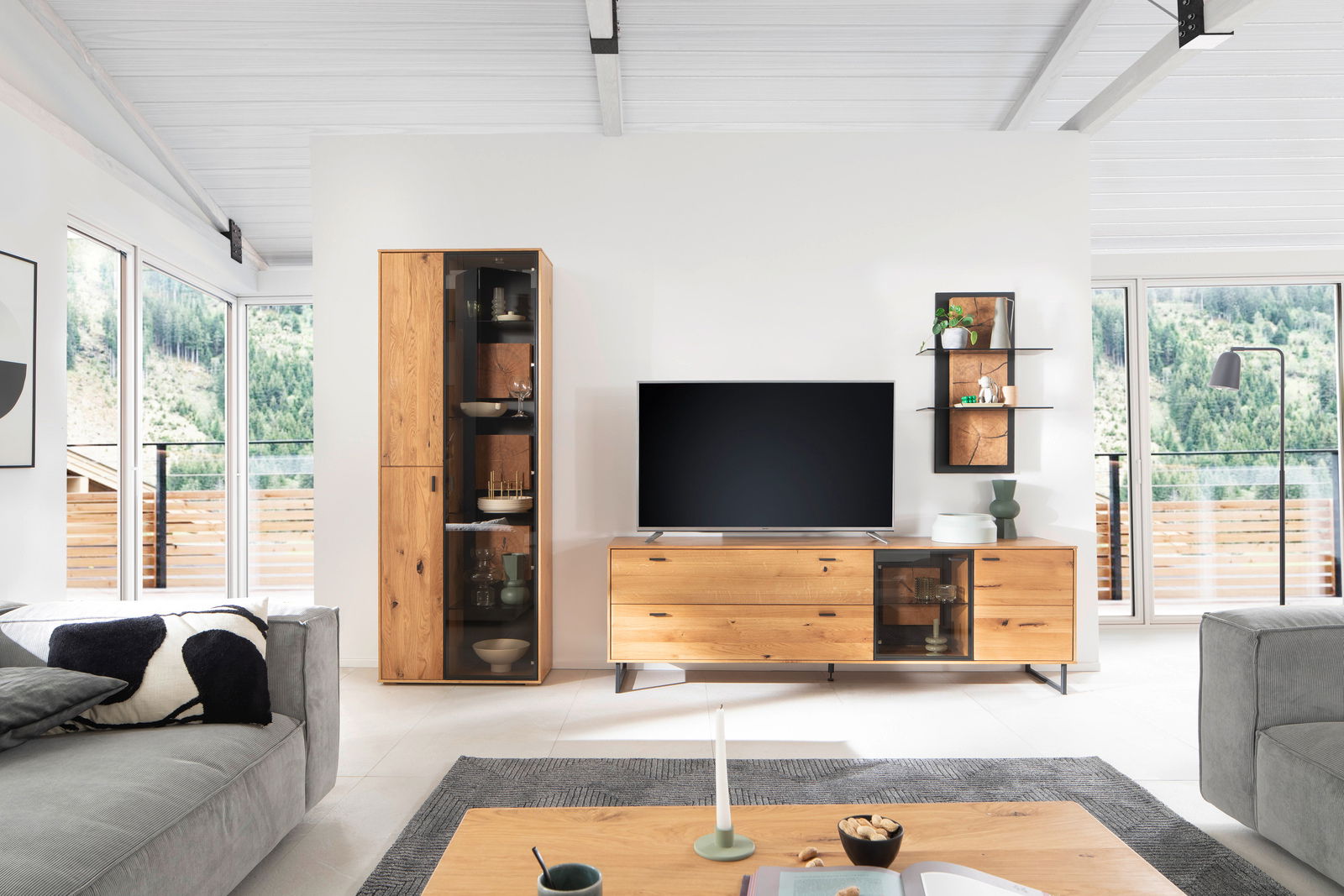 Moderne Wandkombination ASSINI in einem hellen Wohnzimmer mit Blick auf die Berge, bestehend aus einem hohen Schrank, einem TV-Board und einem Wandregal, aus Holz mit Glaselementen.