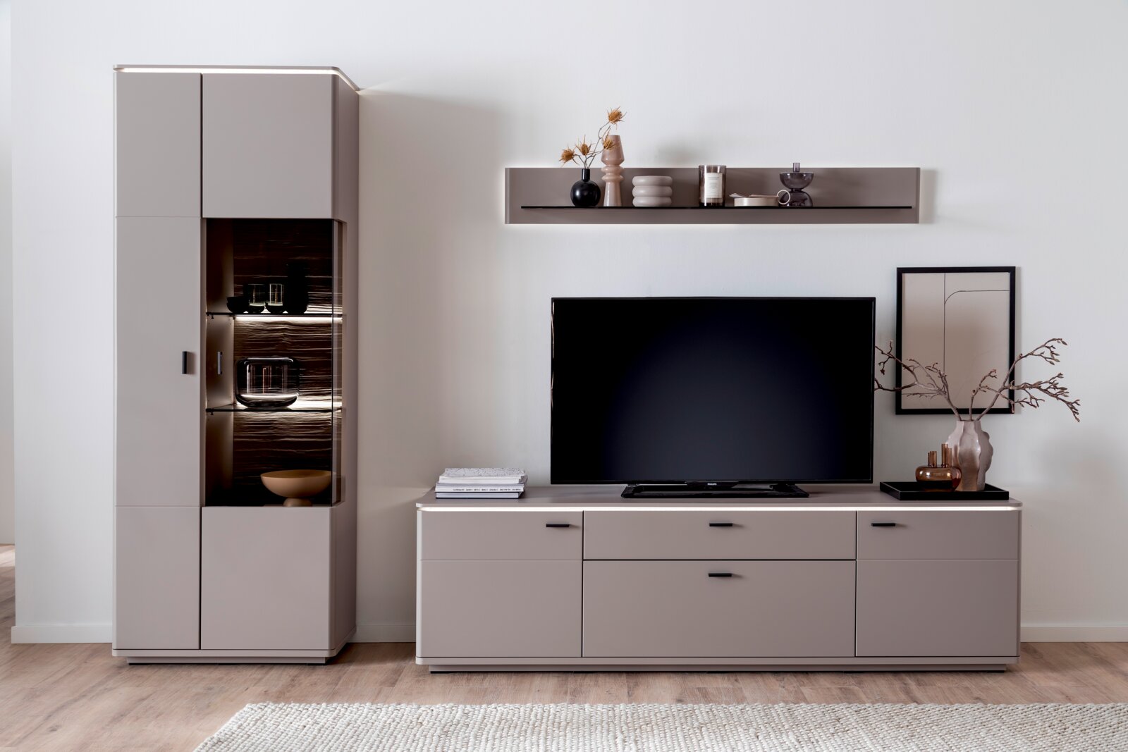 Modernes Wohnzimmer-Entertainment-Set in Taupe mit hoher Vitrine mit beleuchtetem Glasfach, TV-Lowboard und Wandregal, frontal aus Augenhöhe fotografiert.