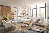 Moderne Wohnzimmer-Einrichtung mit einer hellgrauen Rundecke, zwei Sesseln, einem Couchtisch und großen Fenstern, die viel Licht hereinlassen. Perspektive von vorne auf die Rundecke.