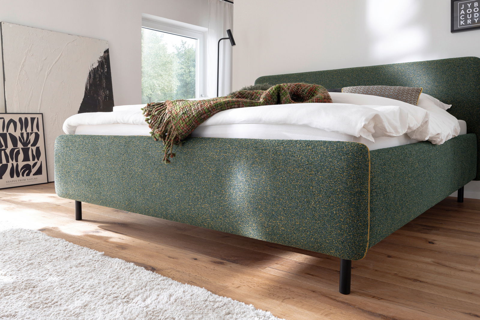 Grünes Polsterbett in einem modernen Schlafzimmer, seitliche Perspektive, mit weißer Bettwäsche und einer gemusterten Decke.