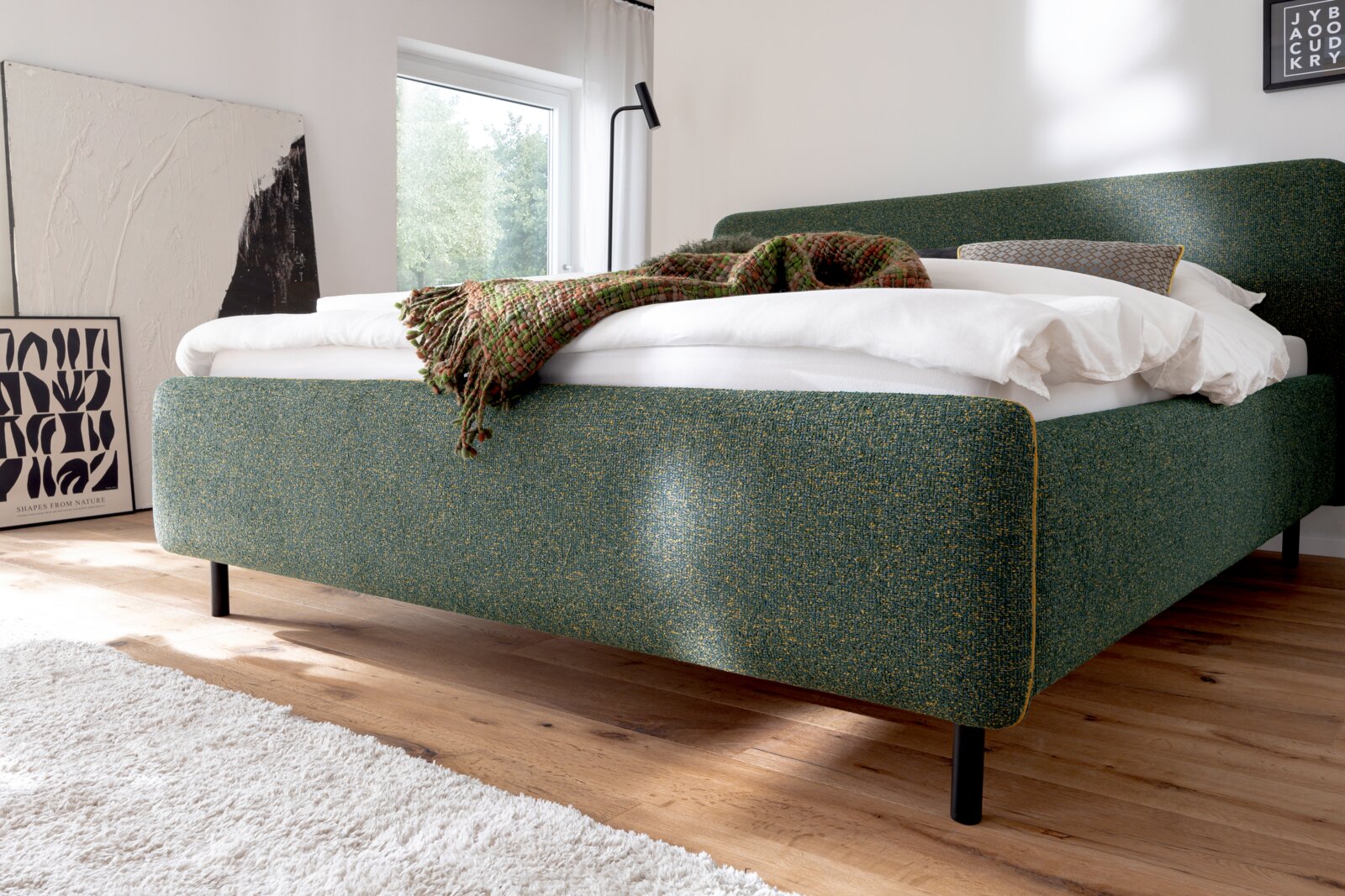 Groen gestoffeerd bed in een moderne slaapkamer, gezien vanaf de zijkant, met wit beddengoed en een gedessineerde deken.