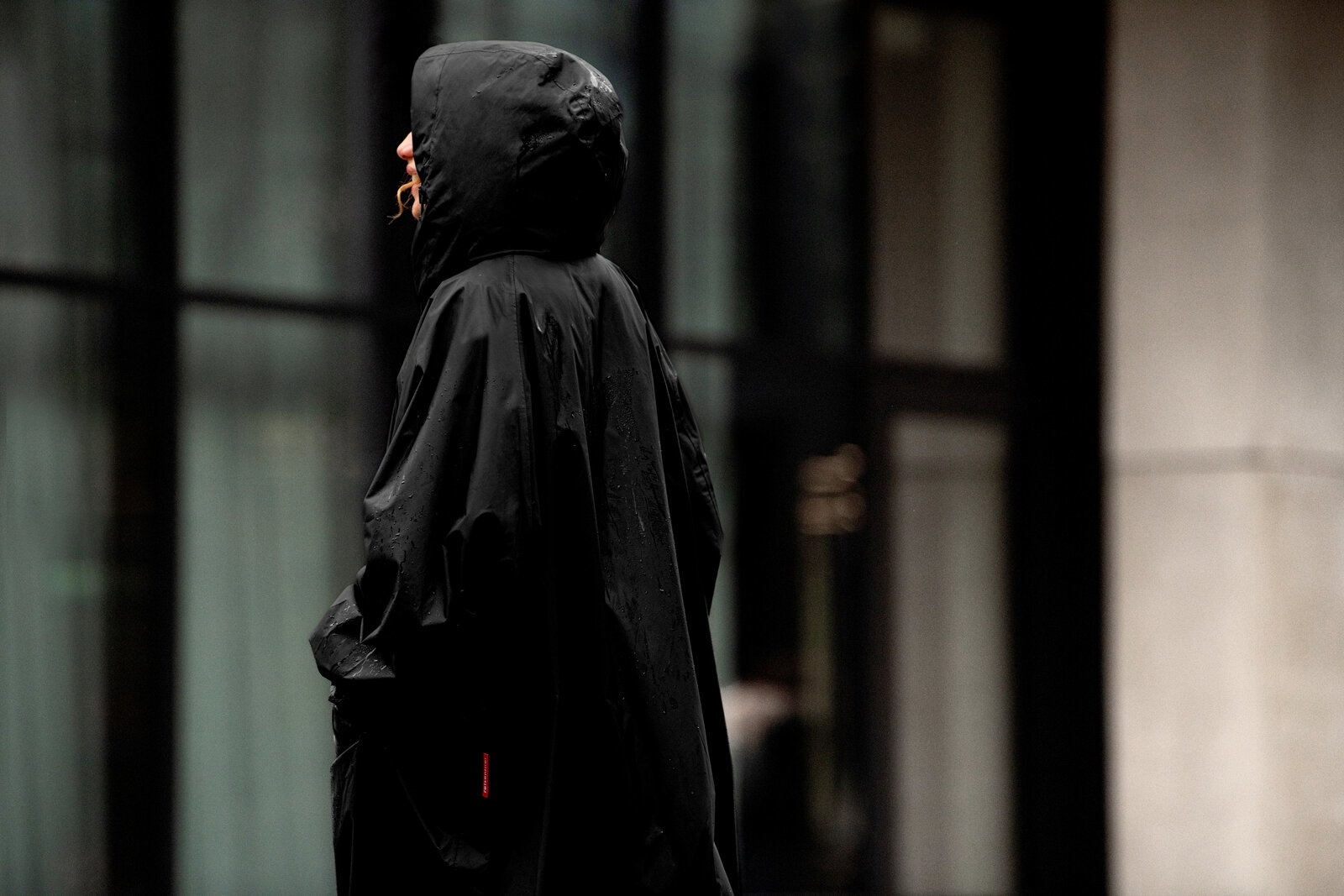 Schwarzer Regenponcho mit Kapuze an einer Person im Regen, seitliche Profilansicht (Seitenperspektive) vor unscharfem urbanem Hintergrund