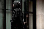 Schwarzer Regenponcho mit Kapuze an einer Person im Regen, seitliche Profilansicht (Seitenperspektive) vor unscharfem urbanem Hintergrund