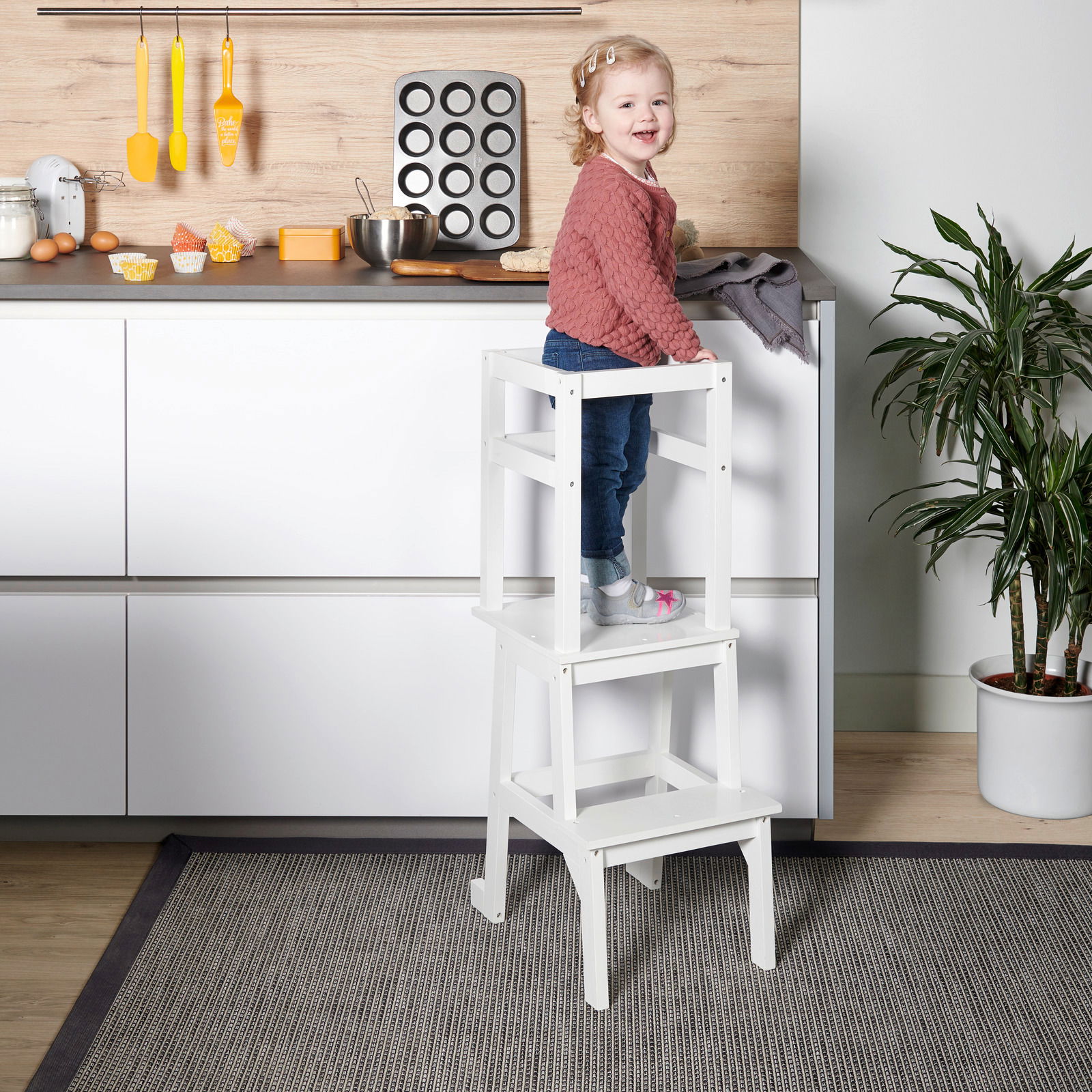Ein weißer Lernturm in einer Küche, auf dem ein kleines Kind steht. Der Lernturm ist von der Seite zu sehen.