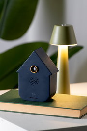 Dunkelblaue Soundbox in Form eines Hauses, frontal auf einem Buch platziert, mit einer Lampe im Hintergrund.