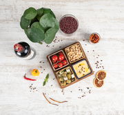 Vogelperspektive auf ein Akazienbrett mit vier Servierschalen, gefüllt mit Tomaten, Nüssen, Oliven und Hummus, umgeben von einer Flasche Wein, einer Pflanze und verschiedenen Gewürzen auf einem hellen Holztisch.