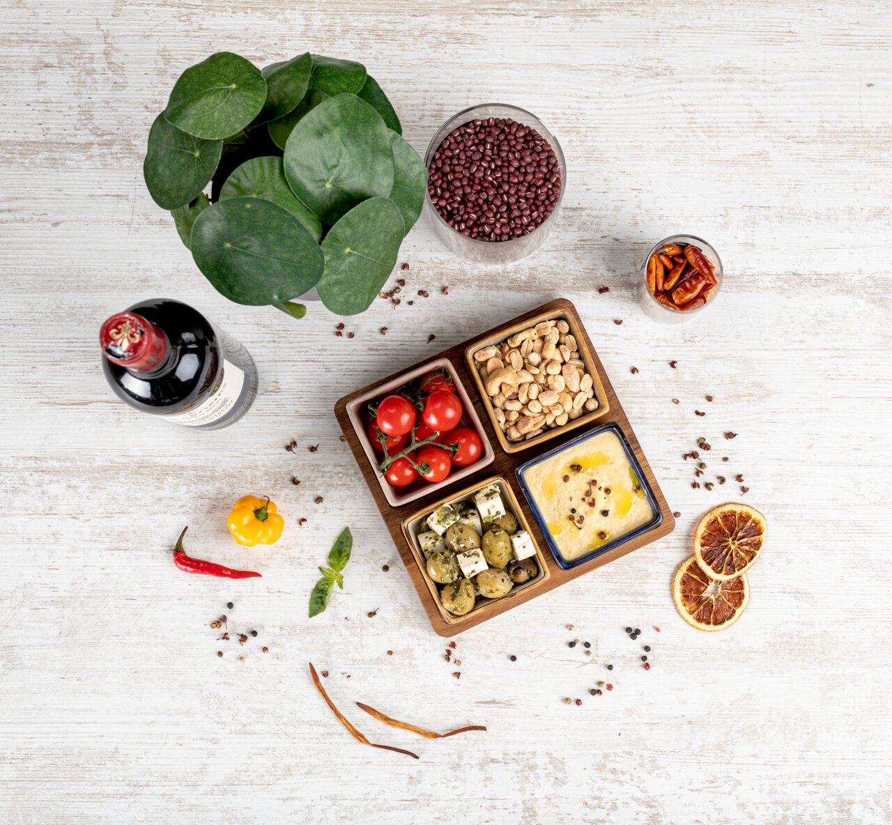 Vogelperspektive auf ein Akazienbrett mit vier Servierschalen, gefüllt mit Tomaten, Nüssen, Oliven und Hummus, umgeben von einer Flasche Wein, einer Pflanze und verschiedenen Gewürzen auf einem hellen Holztisch.