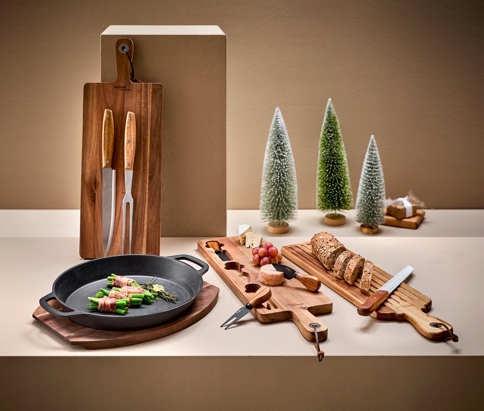 Küchen- und Servier-Set mit Holz-Schneidebrettern, Brotmesser, Käsemessern und gusseiserner Pfanne mit Speckbohnen, dekoriert mit Brot, Käse und Trauben; Perspektive: leicht erhöhte Frontansicht auf einem Tisch.
