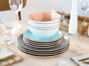 Stapel aus buntem Keramikgeschirr mit Schalen und Tellern in Pastellfarben (rosa, türkis, grau) mit braunem Rand auf gedecktem Tisch, frontale leicht erhöhte Perspektive.