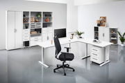 Moderne Büroumgebung mit weißem Schreibtisch, ergonomischem Bürostuhl und mehreren Regalen. Perspektive: Frontalansicht auf den Schreibtisch und die Regale im Hintergrund.