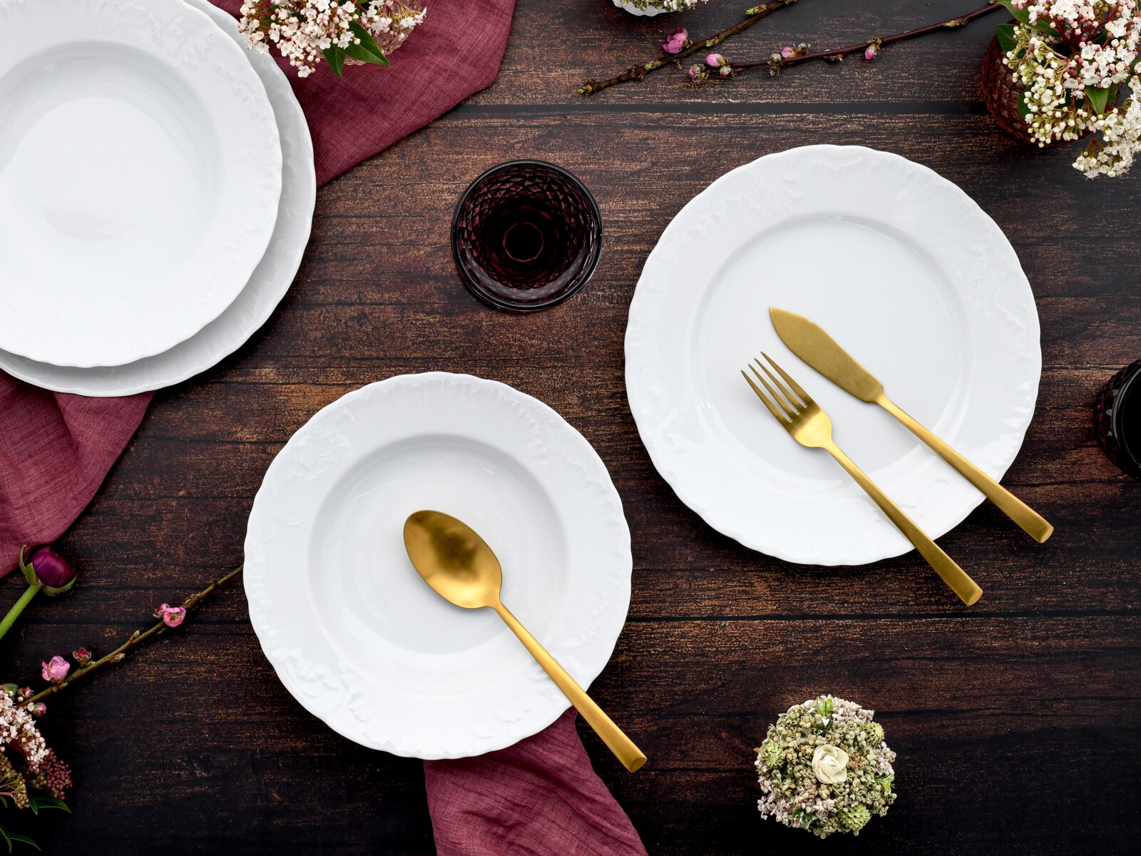 Draufsicht auf elegant gedeckten Tisch mit weißen Porzellantellern mit Reliefrand, goldfarbenem Besteck (Messer, Gabel, Löffel), bordeauxroter Stoffserviette, dunklem Glas und Blumen auf rustikaler Holzplatte