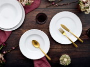 Draufsicht auf elegant gedeckten Tisch mit weißen Porzellantellern mit Reliefrand, goldfarbenem Besteck (Messer, Gabel, Löffel), bordeauxroter Stoffserviette, dunklem Glas und Blumen auf rustikaler Holzplatte