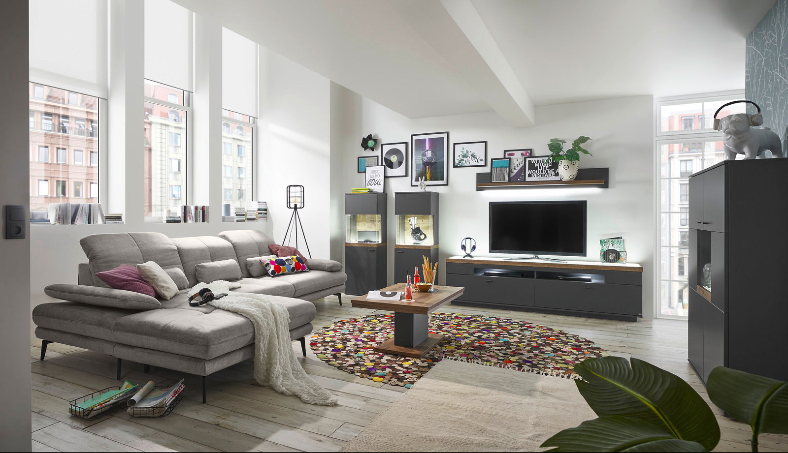 Set one by Musterring Wandpaneel SET ONE LANCASTER Modernes Wohnzimmer mit grauem Sofa, Couchtisch, TV-Lowboard und Vitrinen in einem hellen Raum mit großen Fenstern, Blick von der Seite.