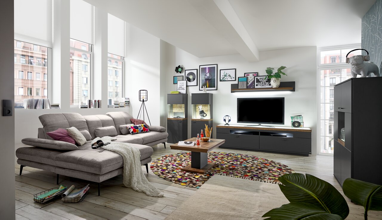 Set one by Musterring Wandpaneel SET ONE LANCASTER Modernes Wohnzimmer mit grauem Sofa, Couchtisch, TV-Lowboard und Vitrinen in einem hellen Raum mit großen Fenstern, Blick von der Seite.