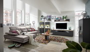 Modernes Wohnzimmer mit grauem Sofa, Couchtisch, TV-Lowboard und Vitrinen in einem hellen Raum mit großen Fenstern, Blick von der Seite.