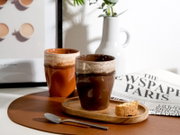 Zwei rustikale Keramikbecher in Braun- und Cremetönen auf einem Holztablett mit Keks, daneben ein Löffel und eine Zeitung; fotografiert in leicht schräger Vorderansicht auf einem Tisch mit braunem Platzset.