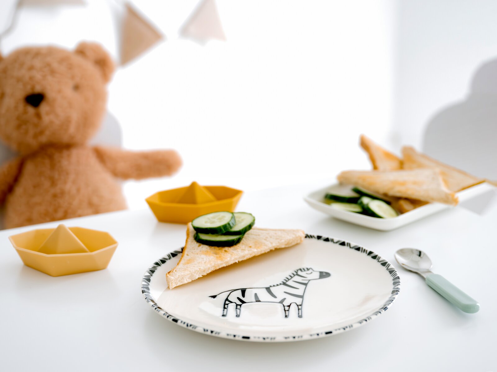 Kinder-Geschirrset mit Zebra-Teller, pastellgrünem Löffel und senfgelben Schälchen in Schiffchenform, aus leicht erhöhter Vorderansicht auf einem gedeckten Tisch präsentiert