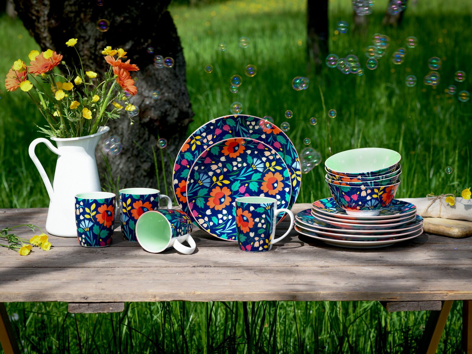 Frontale Ansicht eines bunten Geschirrsets mit floralem Muster (Teller, Schalen und Tassen) auf einem Holztisch im Garten, daneben weiße Kanne mit Blumen und schwebende Seifenblasen