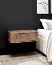 Schwebender Nachttisch aus hellem Holz mit Klappe/Schublade an dunkler Wand neben einem Bett, seitliche Perspektive.