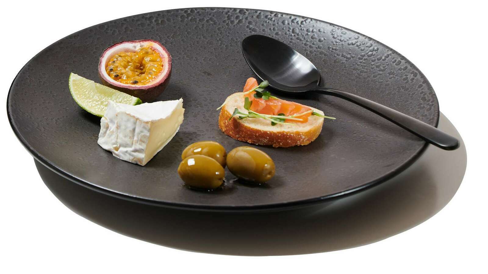 Schwarzer Teller mit strukturiertem Design, darauf eine halbe Passionsfrucht, ein Limettenstück, ein Stück Brie, drei grüne Oliven, ein Stück Baguette mit Lachs und Kresse, und ein schwarzer Löffel. Perspektive von schräg oben.