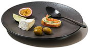 Schwarzer Teller mit strukturiertem Design, darauf eine halbe Passionsfrucht, ein Limettenstück, ein Stück Brie, drei grüne Oliven, ein Stück Baguette mit Lachs und Kresse, und ein schwarzer Löffel. Perspektive von schräg oben.
