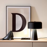 Gerahmtes Poster mit großem Buchstaben „D“ neben einer schwarzen Tischlampe auf einem Sideboard, frontal aus leicht erhöhter Perspektive