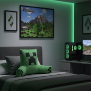Grünes Minecraft Creeper-Kissen als Rückenstütze auf einem grauen Bett, im Hintergrund Gaming-Setup mit grünem LED-Licht und Minecraft-Poster, Perspektive: leicht schräg von vorne.