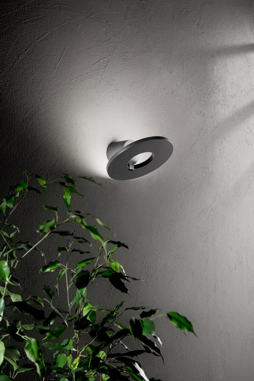 Moderne LED-Außenwandleuchte an einer Wand, seitliche Perspektive, mit Pflanzen im Vordergrund.