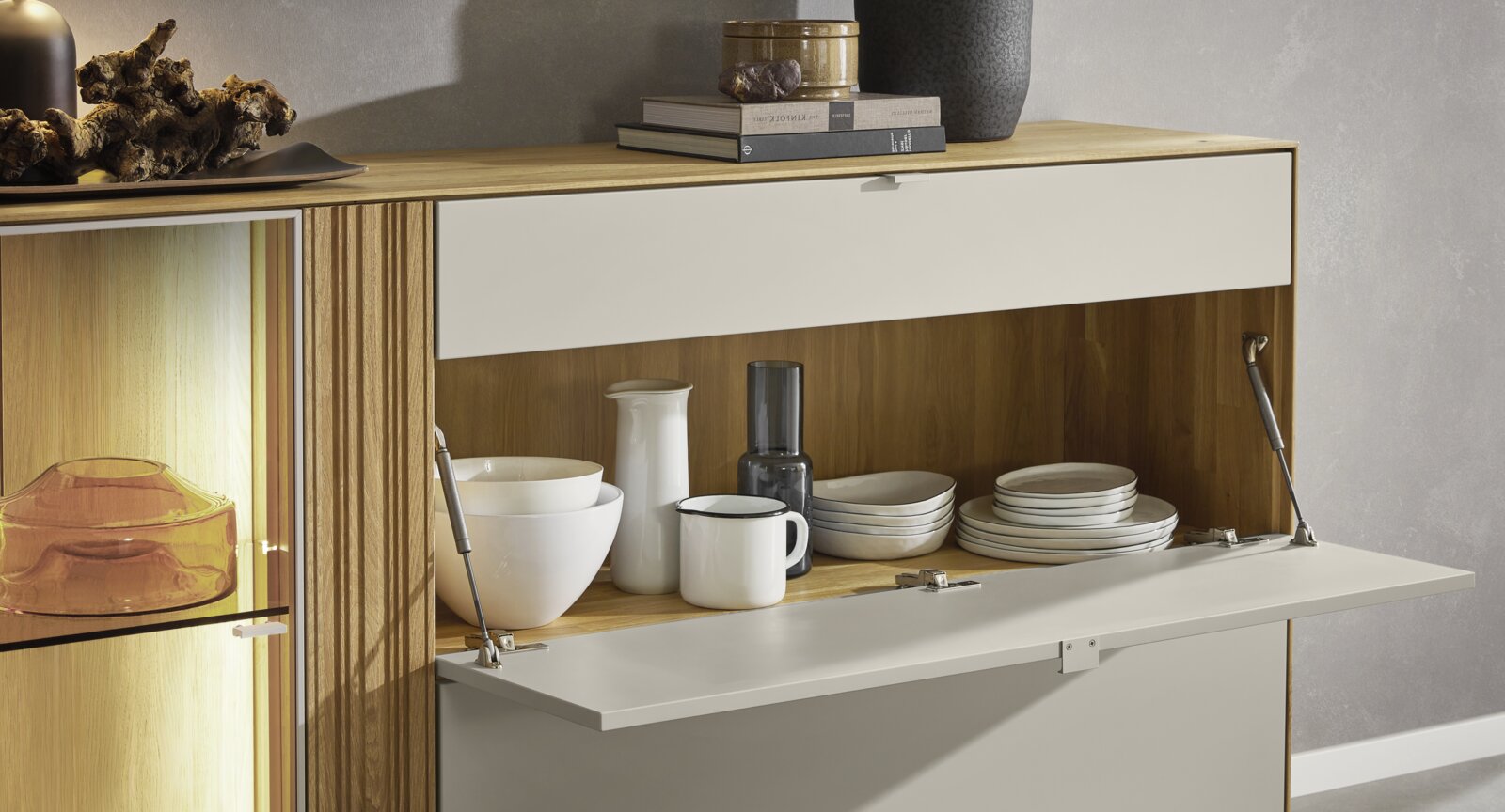 Modernes Sideboard aus Holz mit weißen Klappen, teils geöffnet mit Geschirr und beleuchtetem Glasfach; Frontansicht in leicht schräger Perspektive von vorn rechts.