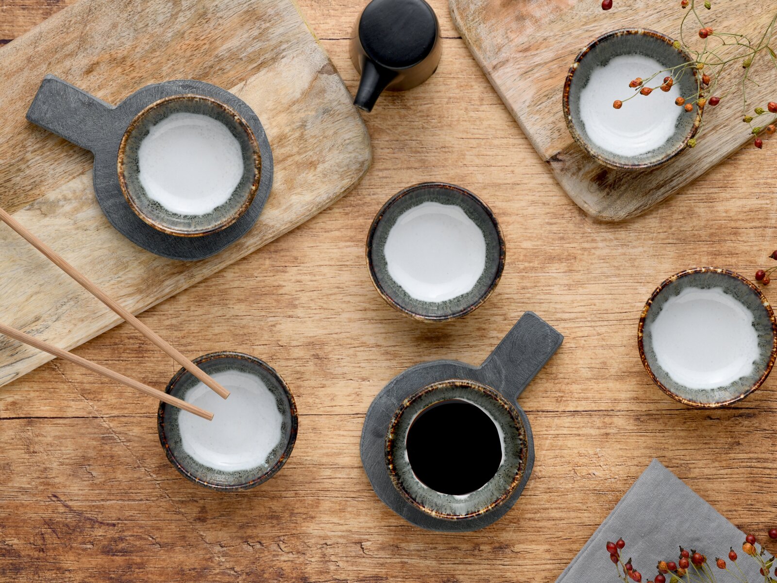 Draufsicht auf mehrere kleine Keramikschalen mit weißer Glasur und braun gesprenkeltem Rand auf einem Holztisch, teils auf dunklen Untersetzern, mit Stäbchen und Dekoelementen.