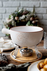 Weiße, gesprenkelte Keramik-Fondue-Schüssel mit Griff auf Metallgestell und Holzuntersetzer, in schräger Vorderansicht auf einem gedeckten Tisch