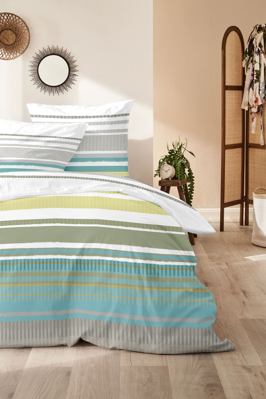 Bettwäsche-Set mit Streifenmuster in Grün-, Blau- und Grautönen, auf einem Bett in einem hellen Raum mit Holzboden und dekorativen Elementen, aus der Vorderansicht fotografiert.