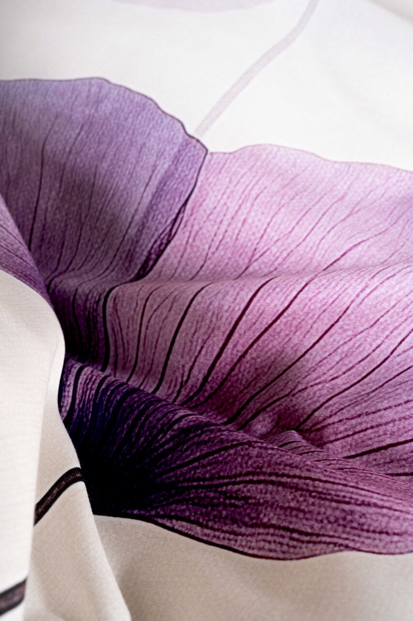 Nahaufnahme aus schräger Draufsicht auf ein weißes Textil mit großem Blütenmotiv in Violett- und Rosatönen, detailreiche Blütenblätter mit feiner Linienstruktur