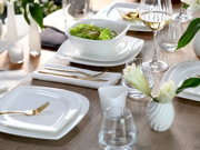 Modern gedeckter Esstisch mit weißem, eckigem Porzellan-Geschirr (Teller und Schale mit Salat), goldfarbenem Besteck und Glaswaren auf Holztisch, fotografiert aus leicht schräger Seitenansicht von oben.