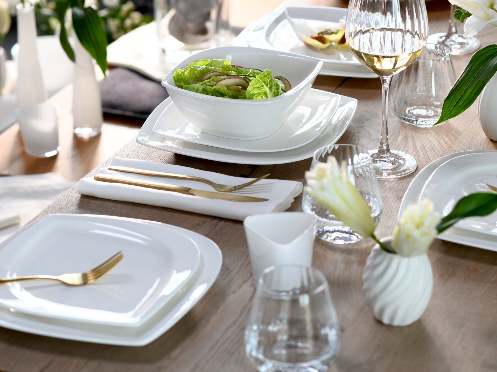 Modern gedeckter Esstisch mit weißem, eckigem Porzellan-Geschirr (Teller und Schale mit Salat), goldfarbenem Besteck und Glaswaren auf Holztisch, fotografiert aus leicht schräger Seitenansicht von oben.