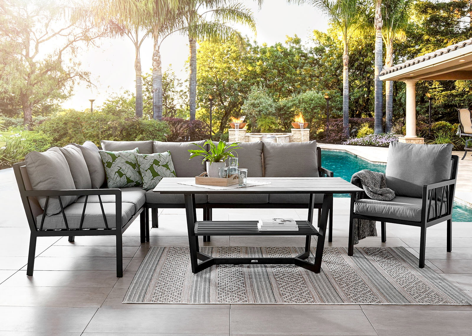 Amatio Gartensessel ZAMORA Gartenset bestehend aus einem Lounge-Sofa, einem Gartensessel und einem Tisch, auf einer Terrasse mit Blick auf einen Pool und umgeben von Palmen.