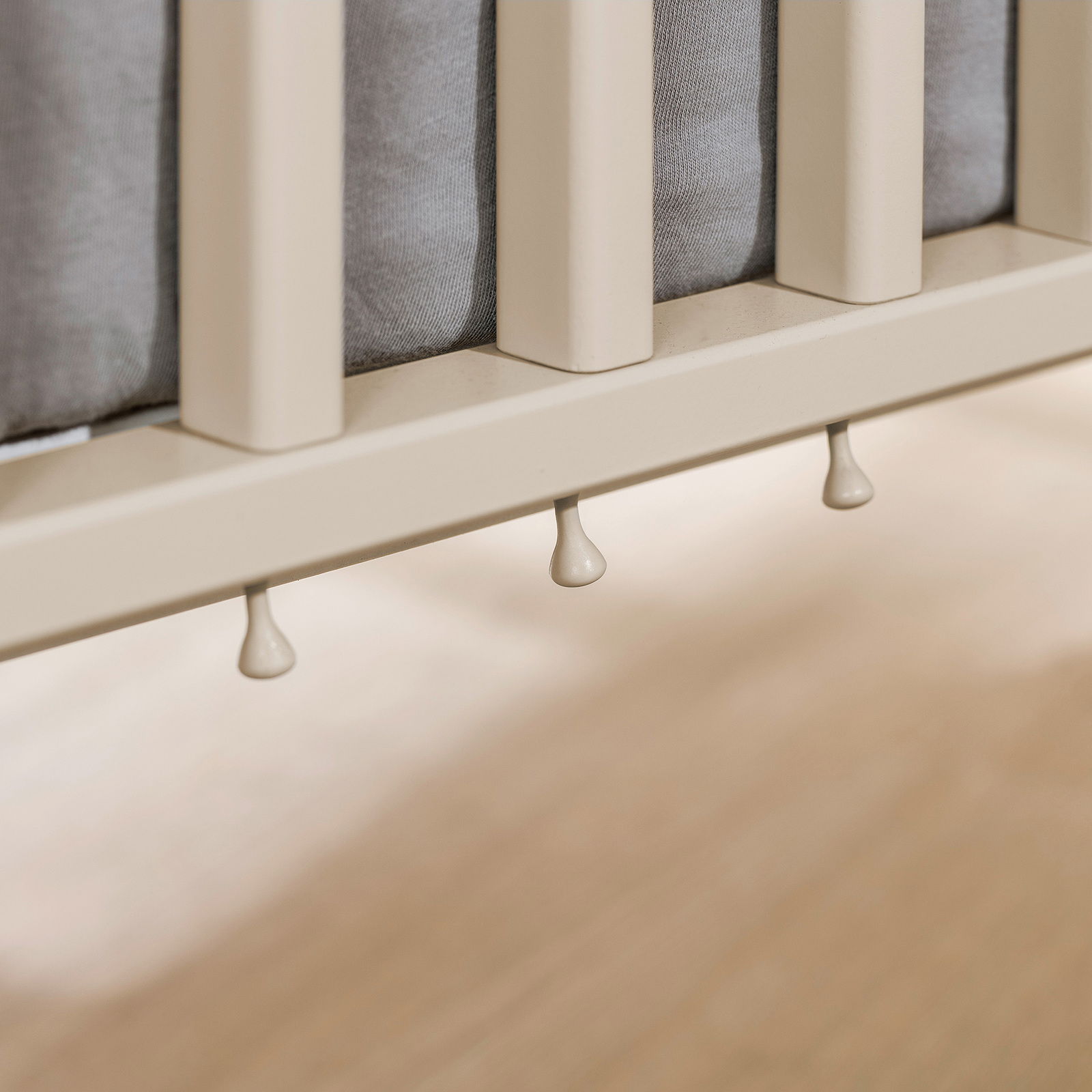 Nahaufnahme eines Babybetts mit cremefarbenen Gitterstäben und grauer Matratze, seitliche Perspektive