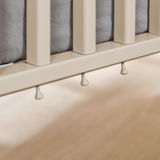 Nahaufnahme eines Babybetts mit cremefarbenen Gitterstäben und grauer Matratze, seitliche Perspektive