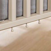 Nahaufnahme eines Babybetts mit cremefarbenen Gitterstäben und grauer Matratze, seitliche Perspektive