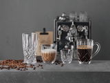 Vier elegante Gläser in verschiedenen Größen auf einer grauen Oberfläche, gefüllt mit Espresso und Kaffee, im Hintergrund eine Espressomaschine. Frontalansicht.