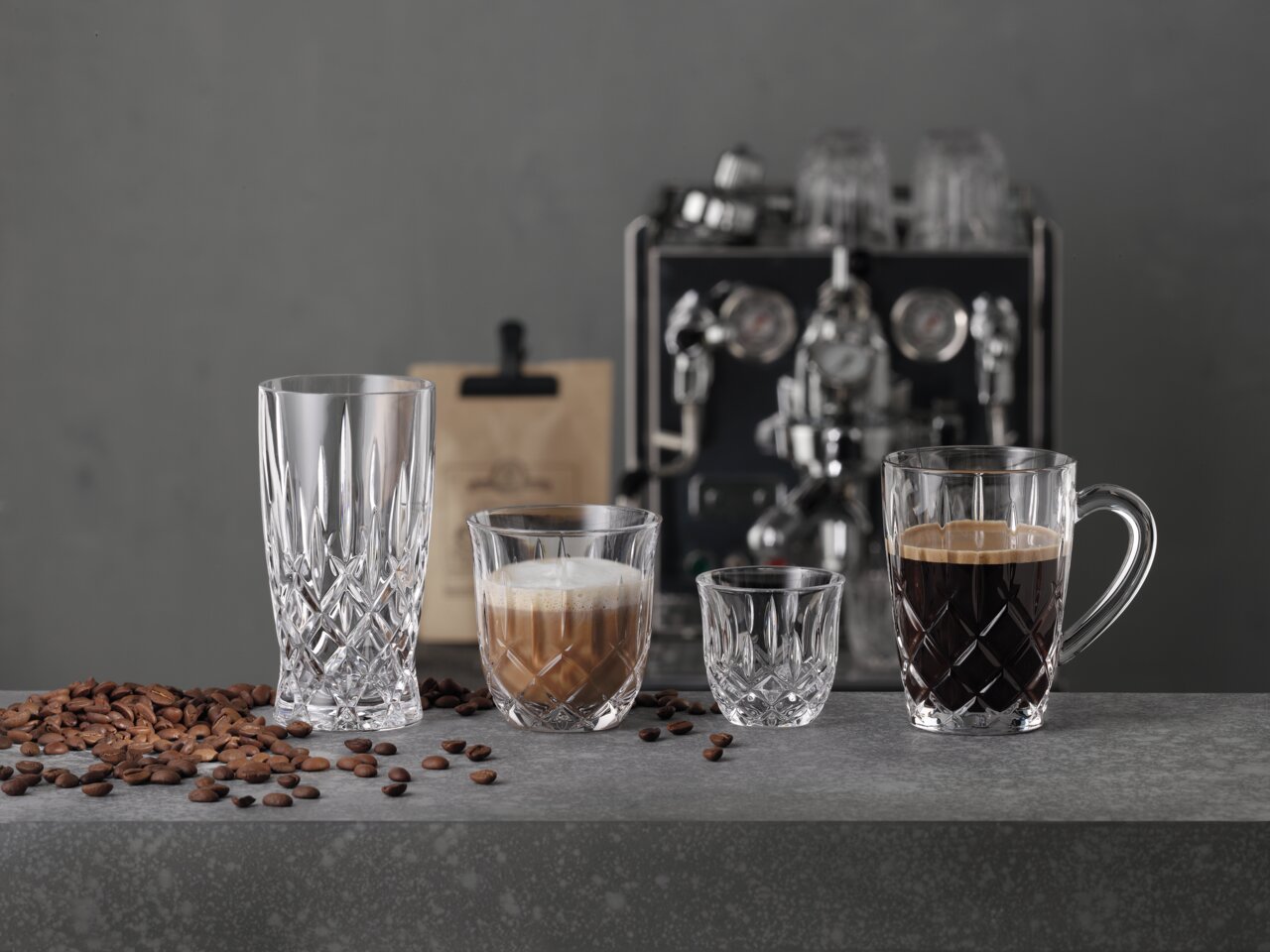 Nachtmann Barista Espresso 2er-Set Vier elegante Gläser in verschiedenen Größen auf einer grauen Oberfläche, gefüllt mit Espresso und Kaffee, im Hintergrund eine Espressomaschine. Frontalansicht.