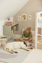 Ein modernes, grünes Wandregal in einem gemütlichen Kinderzimmer, mit einem Kind, das auf dem Boden sitzt und zeichnet, während ein Hund daneben schläft. Perspektive von vorne.
