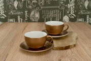 Zwei braune Keramik-Kaffeetassen mit Untertassen auf Holztisch, eine Tasse im Vordergrund und eine im Hintergrund auf einem Holzbrett, fotografiert in leicht erhöhter Frontansicht.