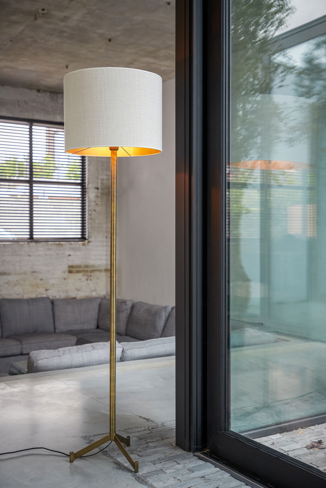 Stehlampe mit weißem zylindrischem Lampenschirm und goldenem Ständer, in einem modernen Wohnzimmer mit Blick auf ein Fenster.